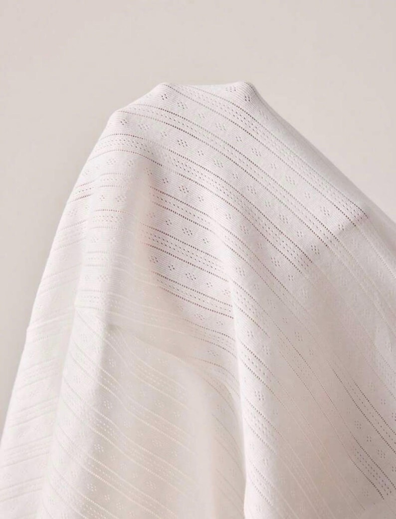 May include: Close-up of white fabric with a delicate, open weave. The fabric has a subtle texture with vertical and diagonal lines, creating a sense of depth and visual interest. The material appears soft and lightweight.