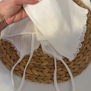 May include: A white baby bonnet with a delicate, textured fabric and scalloped edges. The bonnet has long, white ties for securing. The bonnet is displayed on a woven, circular surface.