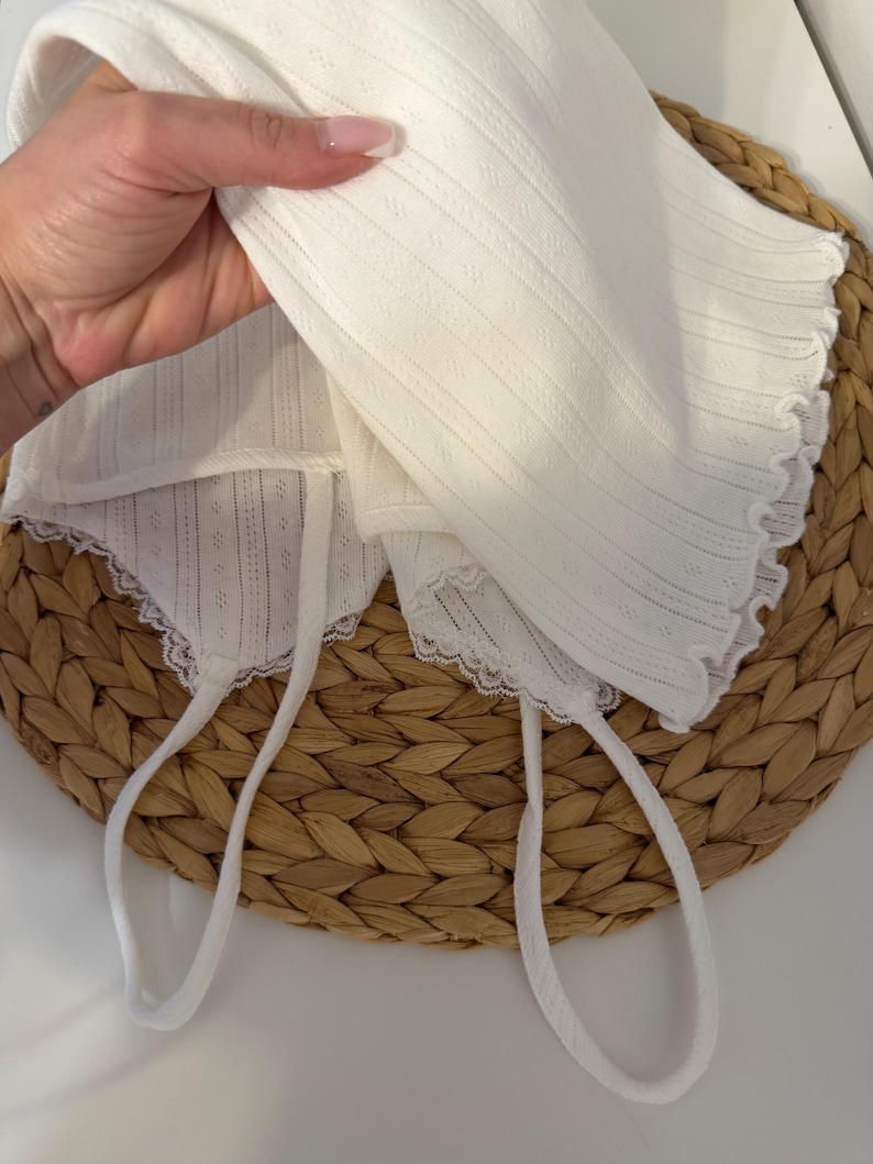 May include: A white baby bonnet with a delicate, textured fabric and scalloped edges. The bonnet has long, white ties for securing. The bonnet is displayed on a woven, circular surface.
