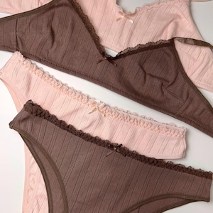 May include: A collection of lingerie in shades of pink and brown. The set includes a bra and a pair of underwear, both with lace trim and small bows. The fabric has a textured, ribbed pattern. The Luna Intimates logo is visible.