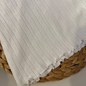 May include: White, textured fabric with vertical lines and small diamond-shaped perforations. The edge of the fabric has a scalloped trim. The fabric is likely a clothing item, possibly a top or a dress.