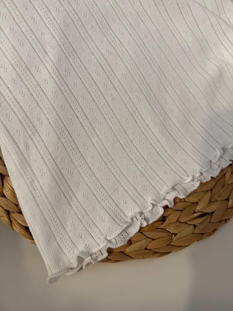 May include: White, textured fabric with vertical lines and small diamond-shaped perforations. The edge of the fabric has a scalloped trim. The fabric is likely a clothing item, possibly a top or a dress.