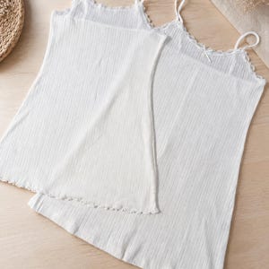 May include: Two white ribbed camisole tops with spaghetti straps and scalloped edges. The tops are made of a soft, lightweight fabric and are suitable for layering or wearing on their own. The camisoles are a versatile addition to any wardrobe.