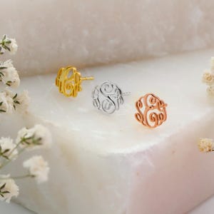 May include: Three monogram stud earrings in gold, silver, and rose gold. Each earring features a delicate, swirling letter design. The earrings are displayed on a white marble surface, with sprigs of white flowers in the foreground.