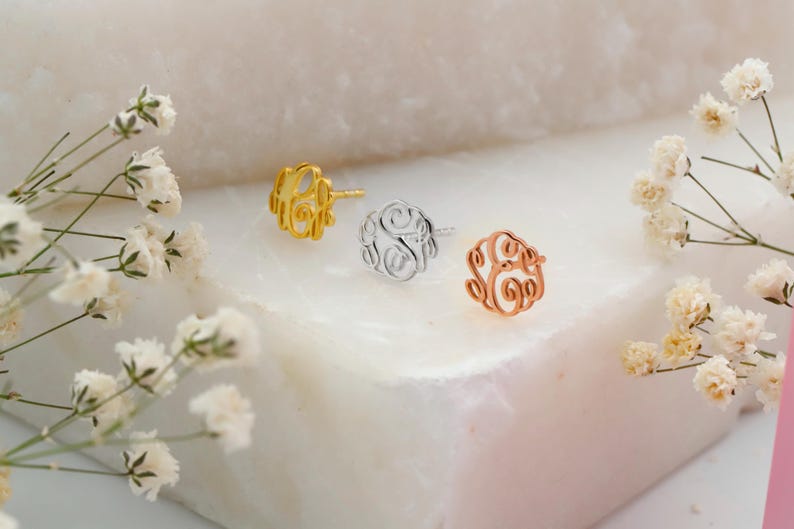 May include: Three monogram stud earrings in gold, silver, and rose gold. Each earring features a delicate, swirling letter design. The earrings are displayed on a white marble surface, with sprigs of white flowers in the foreground.
