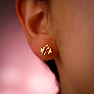 May include: A gold-colored monogram earring is displayed on an ear. The earring features a script monogram design, likely representing initials. The earring is a stud style, suitable for everyday wear. The background is blurred, focusing attention on the jewelry.