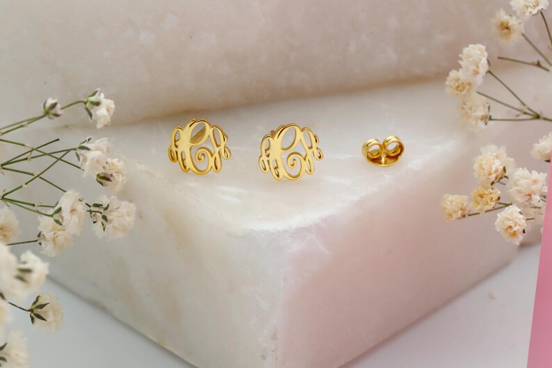 May include: A pair of gold-colored monogram earrings with a matching earring back. The earrings are displayed on a white and pink surface, with white flowers in the background. The earrings have a delicate, openwork design.