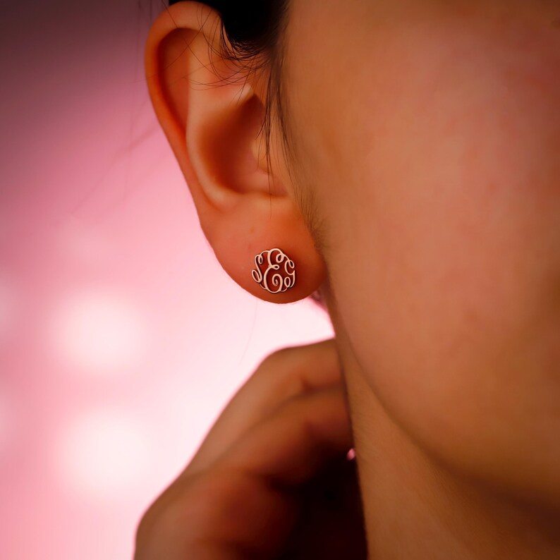 May include: A close-up of a gold-colored monogram earring. The earring is a small, circular stud with an ornate, swirling design. The earring is worn on the ear of a person, with a soft pink background.
