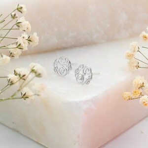 May include: A pair of silver-colored stud earrings with an openwork floral design. The earrings are displayed on a white surface, with sprigs of white and yellow flowers in the foreground. A pink geometric shape is in the background.