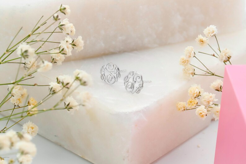 May include: A pair of silver-colored stud earrings with an openwork floral design. The earrings are displayed on a white surface, with sprigs of white and yellow flowers in the foreground. A pink geometric shape is in the background.
