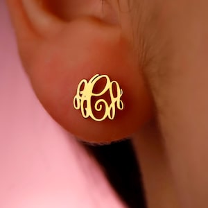 May include: A gold-colored monogram earring with a three-letter design. The earring is circular and features an elegant, cursive font. The earring is shown on an ear against a pink background.