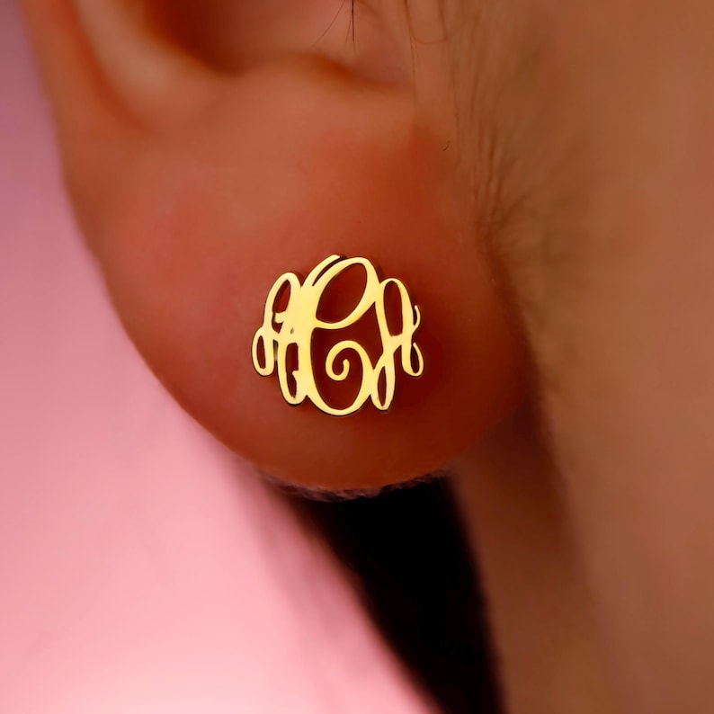 May include: A gold-colored monogram earring with a three-letter design. The earring is circular and features an elegant, cursive font. The earring is shown on an ear against a pink background.