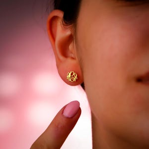 May include: A close-up of a gold-colored monogram stud earring on an ear. The earring features an elegant, intertwined design. The background is a soft gradient of pink and purple.