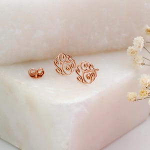 May include: Rose gold filigree earrings and earring backings are displayed on a white marble surface. The earrings have a circular design with an open, swirling pattern. White flowers and a pink geometric shape are in the background.