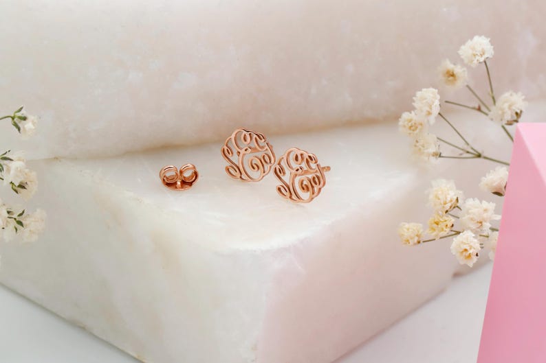 May include: Rose gold filigree earrings and earring backings are displayed on a white marble surface. The earrings have a circular design with an open, swirling pattern. White flowers and a pink geometric shape are in the background.