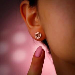 May include: A close-up of a gold-colored monogram earring. The earring is a circular design with an ornate, swirling letter "S" in the center, surrounded by decorative flourishes. The earring is in focus, set against a soft, blurred pink background.