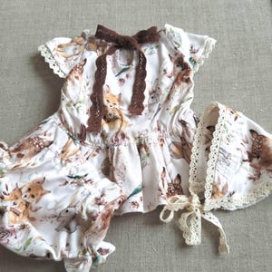 May include: A white baby outfit set with a woodland animal print, including a dress, bloomers, and bonnet. The dress features a brown lace bow and trim. The bonnet has a lace trim and tie. The set is designed for infants.