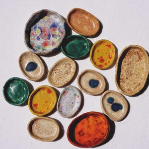 Pode incluir: Uma coleção de pequenos pratos de cerâmica moldados à mão, de formas irregulares e vários acabamentos. O conjunto inclui bandejas de cor bege salpicado, verde sólido, amarelo com manchas laranja, laranja vibrante e esmaltes multicoloridos.