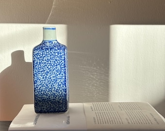 Op de afbeelding: Een rechthoekige blauw-witte keramische fles met een bloemmotief. De fles staat op een boek met de titel "Designing Design" van Kenya Hara.