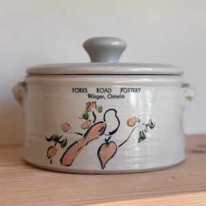 Puede incluir: Olla de cerámica beige con tapa y asas laterales, con un diseño pintado a mano de un pájaro y follaje. El texto "FORKS ROAD POTTERY Winger, Ontario" está impreso en el lateral. La tapa es gris.