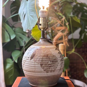 Puede incluir: Lámpara de mesa de cerámica con base redonda y texturizada en tonos marrones y beige. La lámpara presenta un cuello de latón y una bombilla brillante de bajo consumo. La lámpara está colocada sobre una superficie de madera.