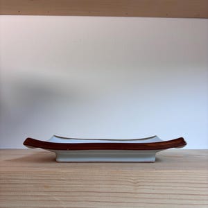 May include: A rectangular white ceramic dish with a brown rim and accents. The dish has a slightly curved shape and is displayed on a wooden surface. The design is simple and elegant, suitable for serving food or as a decorative item.