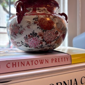 Könnte beinhalten: Eine runde, weiße Keramikvase mit roten Akzenten und Griffen. Die Vase ist mit Blumen- und Vogelmotiven in Rosa-, Rot- und Goldtönen verziert. Die Vase steht auf einem Stapel Bücher, darunter eines mit dem Titel "Chinatown Pretty."