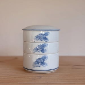 May include: A stackable white ceramic container with a lid, featuring a blue floral design. The container is composed of three sections, each adorned with a blue flower motif. A blue line accents the base and rim, creating a cohesive aesthetic. The container is on a wooden surface.