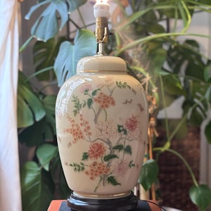 Peut inclure: Lampe en céramique crème avec un motif floral de fleurs roses, oranges et blanches et de feuilles vertes. La lampe a une base noire et un col doré. Une ampoule allumée est au sommet de la lampe.