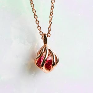 May include: A rose gold necklace with a pendant featuring a red gemstone encased in a cage-like design. The pendant hangs from a delicate chain, creating an elegant and eye-catching piece of jewelry.