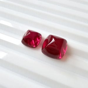 May include: Two faceted, square-shaped, deep red glass beads. The beads have a glossy finish and are set against a white, ridged surface. The light reflects off the facets, creating highlights and shadows.