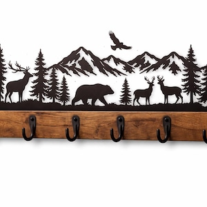 Può includere: Un portachiavi rustico con una silhouette in metallo nero di una scena di montagna con alberi, cervi, un orso e un'aquila. Il design è montato su una tavola di legno macchiato con sei ganci in metallo nero per chiavi.