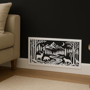 Può includere: Un ritaglio di carta bianca di una scena di montagna con alberi, animali e un fiume. L'opera d'arte è esposta contro un muro nero, con un divano beige e un tavolo di legno sullo sfondo. La scena comprende un cervo, un orso e un lupo.
