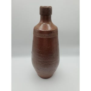 May include: A brown ceramic bottle with a narrow neck and a rounded body. The bottle has a textured surface and horizontal lines near the top of the body. The bottle is likely a decorative piece or a container for liquids.