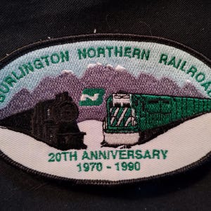 May include: An embroidered patch featuring the Burlington Northern Railroad logo. The patch has an oval shape with a white border. The design includes a black train, a green train, mountains, and the text "20th Anniversary 1970-1990". The text "Burlington Northern Railroad" is arched across the top.