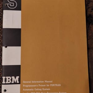 Puede incluir: Un manual de información general de IBM titulado Programmer's Primer for FORTRAN Automatic Coding System for the IBM 704 Data Processing System. La cubierta es de color amarillo mostaza con un lomo vertical en blanco y negro con la letra S.