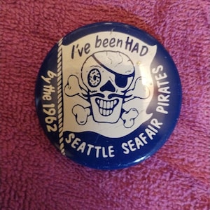 May include: A round, blue button with white text and a pirate skull graphic. The text reads "I've been HAD by the 1962 Seattle Seafair Pirates."