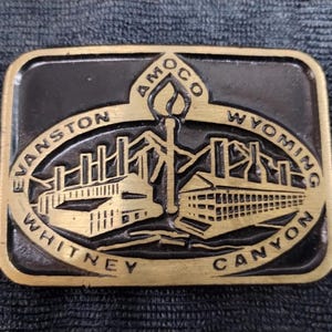 Puede incluir: Hebilla de cinturón rectangular con fondo negro y diseño en relieve dorado. El diseño incluye las palabras "Evanston Wyoming Whitney Canyon" y "Amoco" que rodean una imagen central de una fábrica y una antorcha.