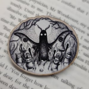 May include: A round wooden coaster with a black and white illustration of a moth with a spooky face and four smaller figures surrounding it. The text "You forget how long I have been here." is printed on the coaster.