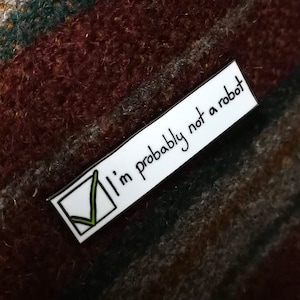 Könnte beinhalten: Eine weiße Emaille-Anstecknadel mit einem grünen Häkchen in einem Kästchen und dem Text "Ich bin wahrscheinlich kein Roboter".