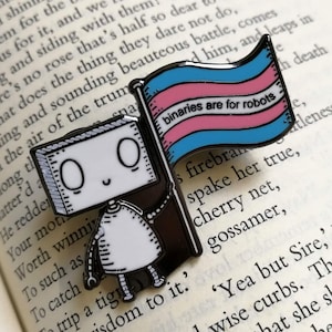 Op de afbeelding: Zwart-witte emaille pin met een robot die een vlag vasthoudt met de transgender pride kleuren en de tekst "binaries are for robots". De pin is geplaatst op de pagina's van een open boek.
