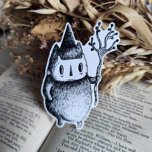 May include: Black and white illustration of a cat wearing a witch's hat and holding a stick. The cat has a mask over its eyes and a small, pointed hat. The cat is standing on a white background.