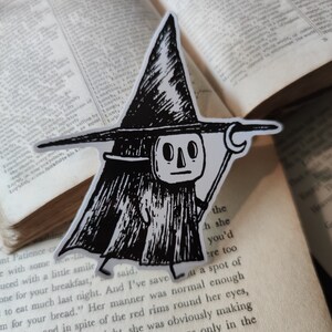May include: A black and white sticker featuring a cartoon character in a tall hat and cloak, holding a staff. The sticker is placed on an open book with printed text. The character has a skull-like face and is walking.
