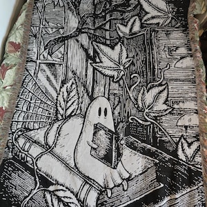 May include: Black and white woven throw blanket with a ghost reading a book design. The illustration includes leaves, books, and a window. The blanket has a fringed edge, suitable for a bed or sofa.