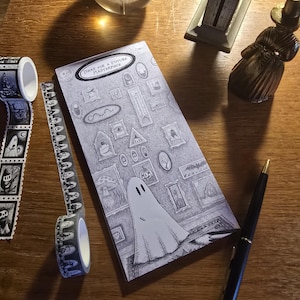 Könnte beinhalten: Ein rechteckiger Notizblock mit einer schwarz-weißen Illustration eines Geistes in einem Raum mit gerahmten Bildern an der Wand und dem Text IDEAS FOR A FUTURE MASTERPIECE, begleitet von zwei Rollen gemustertem Washi-Tape und einem schwarzen Stift.