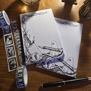 以下が含まれることがあります： ゴースト、鉛筆、棘のある蔓が黒インクで描かれた2冊の白い長方形のメモ帳。その横には、ゴースト柄の装飾テープ2巻と、ゴールドのトリムが付いた黒い万年筆が置かれています。