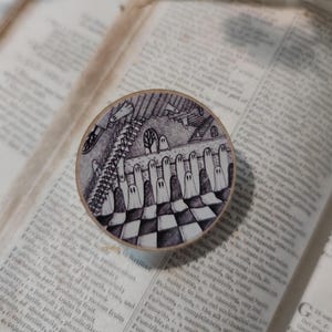 May include: A round wooden coaster with a black and white checkered pattern. The coaster features a black and white illustration of a group of ghosts standing in a line on a staircase.