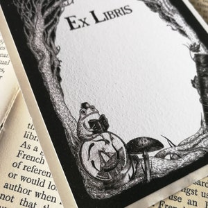 Op de afbeelding: Een zwart-witte exlibris met de tekst "Ex Libris" in een decoratief lettertype. Een kikker met een hoed en een boek zit op een paddenstoel onder een boom.