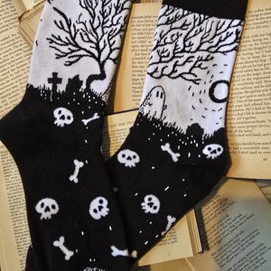 May include: Black and white socks with a spooky graveyard design. The socks have a black top and toe, with a white background featuring a tree, tombstones, skulls, bones, and a ghost. The text "@THISISTOI" is printed on the socks.