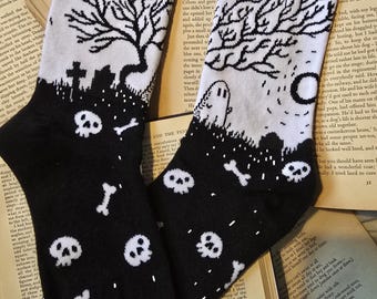May include: Black and white socks with a spooky graveyard design. The socks feature a black top and toe, with a white background showcasing a tree, tombstones, skulls, bones, and a ghost. The text "@THISISTOI" is printed on the socks.